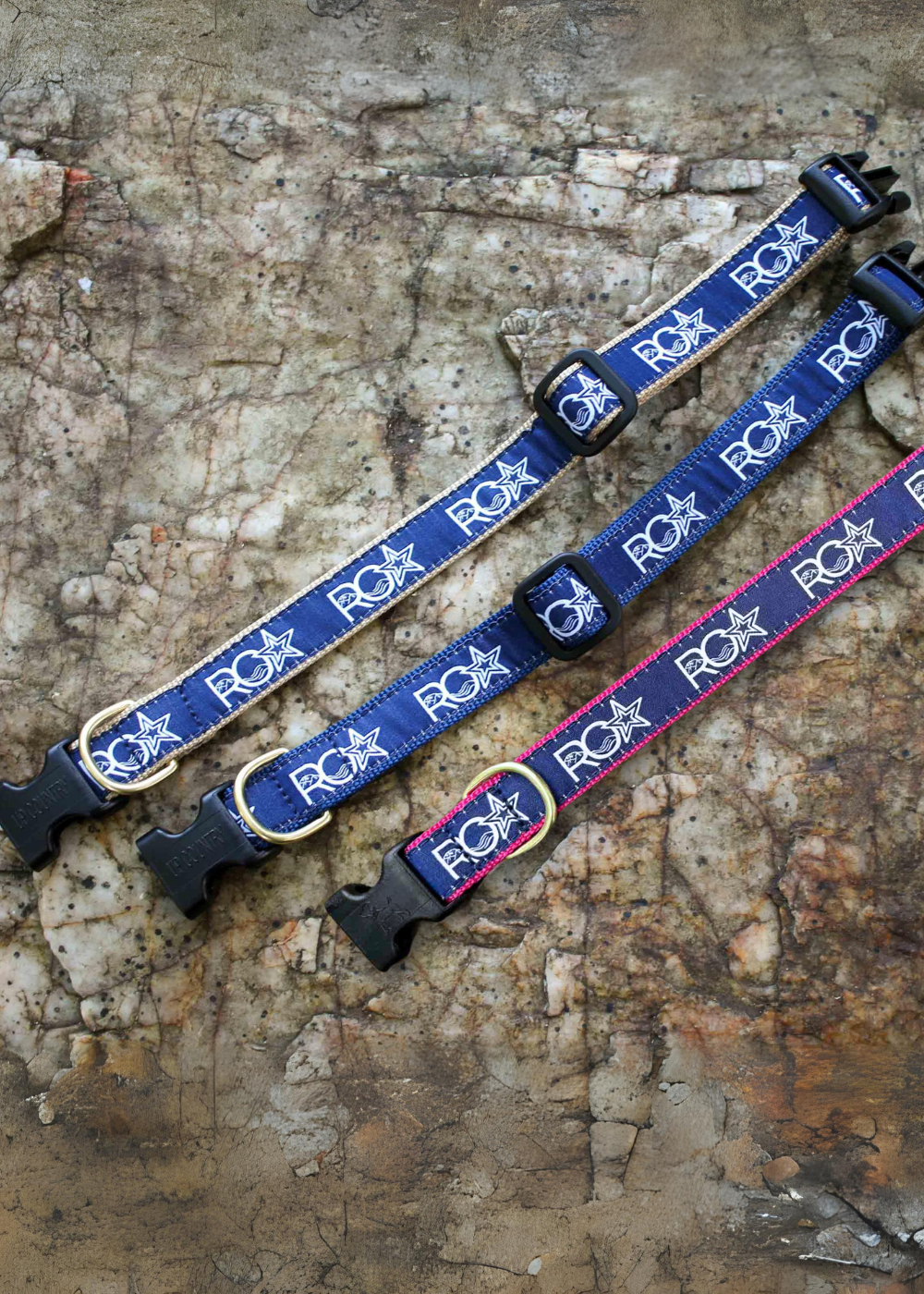 Three ROA-branded dog collars laid out on a natural rock surface. The collars feature navy blue webbing with white ROA logos, each with a brass D-ring and black plastic quick-release buckle. One collar has pink stitching, another has gold stitching, and the third has navy stitching.