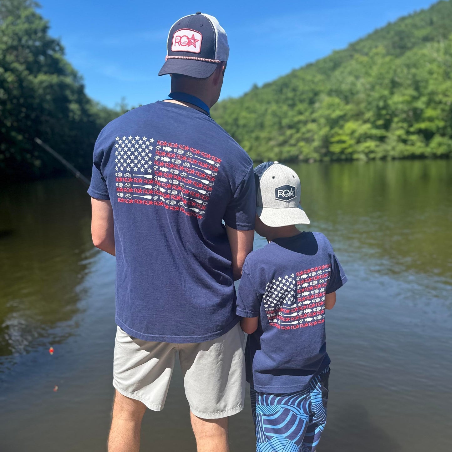 A father and son wearing matching ROA Lifestyle American Flag T-shirts, standing by a lake with their backs to the camera. The shirts feature a unique American flag design on the back, and both are wearing ROA branded hats while enjoying an outdoor adventure together.
