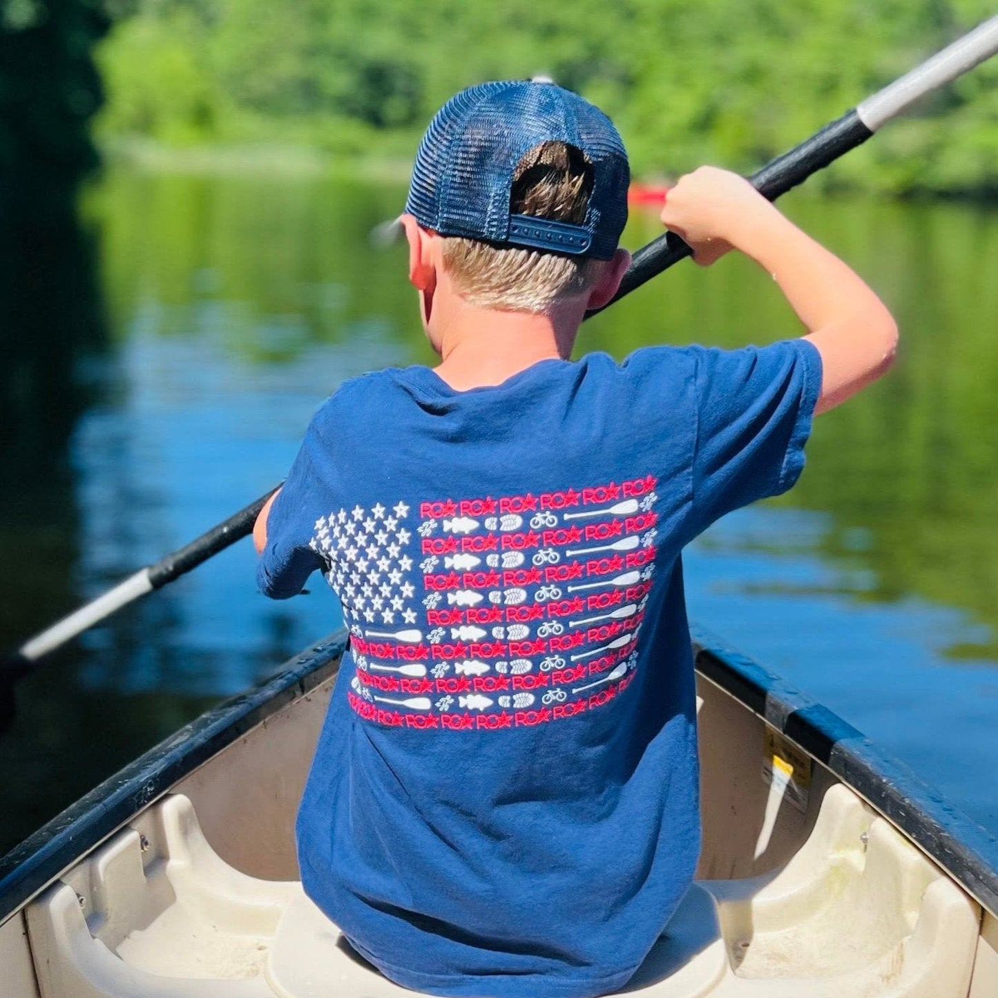 A child wearing the ROA Lifestyle American Flag T-shirt paddles a canoe on a calm lake. The shirt features a unique American flag design on the back, and the child wears a blue cap while enjoying the outdoor adventure.
