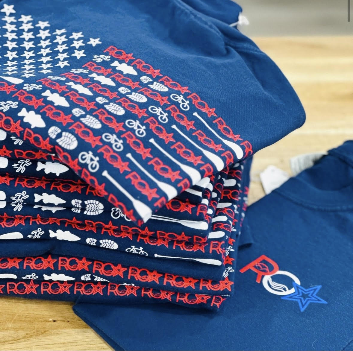 A stack of ROA Lifestyle American Flag T-shirts folded neatly, showcasing the detailed red, white, and blue flag design on the back. The shirts are displayed on a wooden surface, highlighting the unique graphic elements and local craftsmanship.