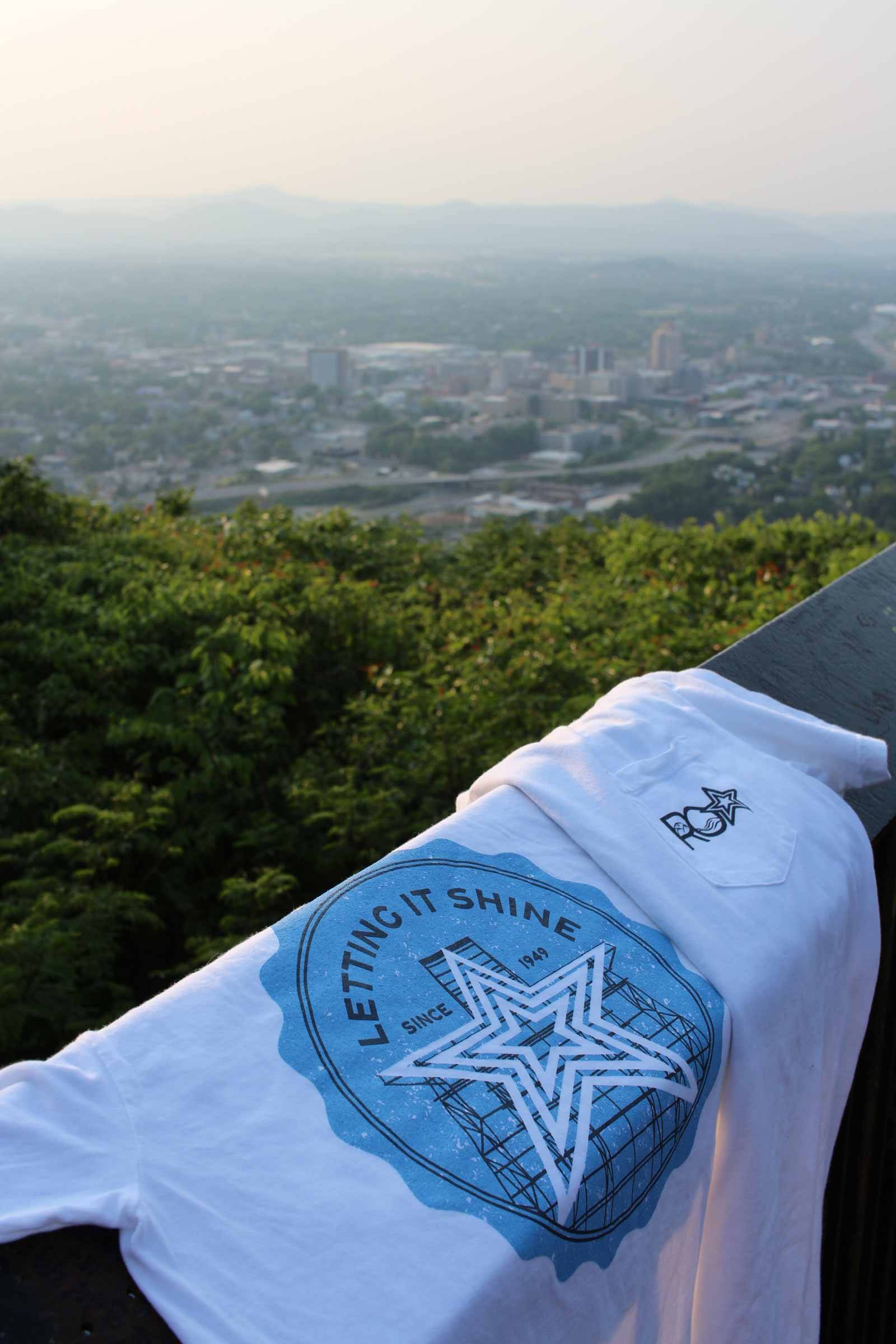 A "Letting It Shine" Roanoke Lifestyle t-shirt draped over a railing, overlooking a scenic view of downtown Roanoke. The t-shirt's large blue graphic on the back features the Mill Mountain Star and the text "Letting It Shine Since 1949." The front pocket displays a small "ROA" logo, celebrating the iconic symbol of the city.