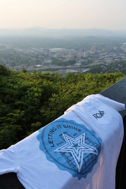 A "Letting It Shine" Roanoke Lifestyle t-shirt draped over a railing, overlooking a scenic view of downtown Roanoke. The t-shirt's large blue graphic on the back features the Mill Mountain Star and the text "Letting It Shine Since 1949." The front pocket displays a small "ROA" logo, celebrating the iconic symbol of the city.