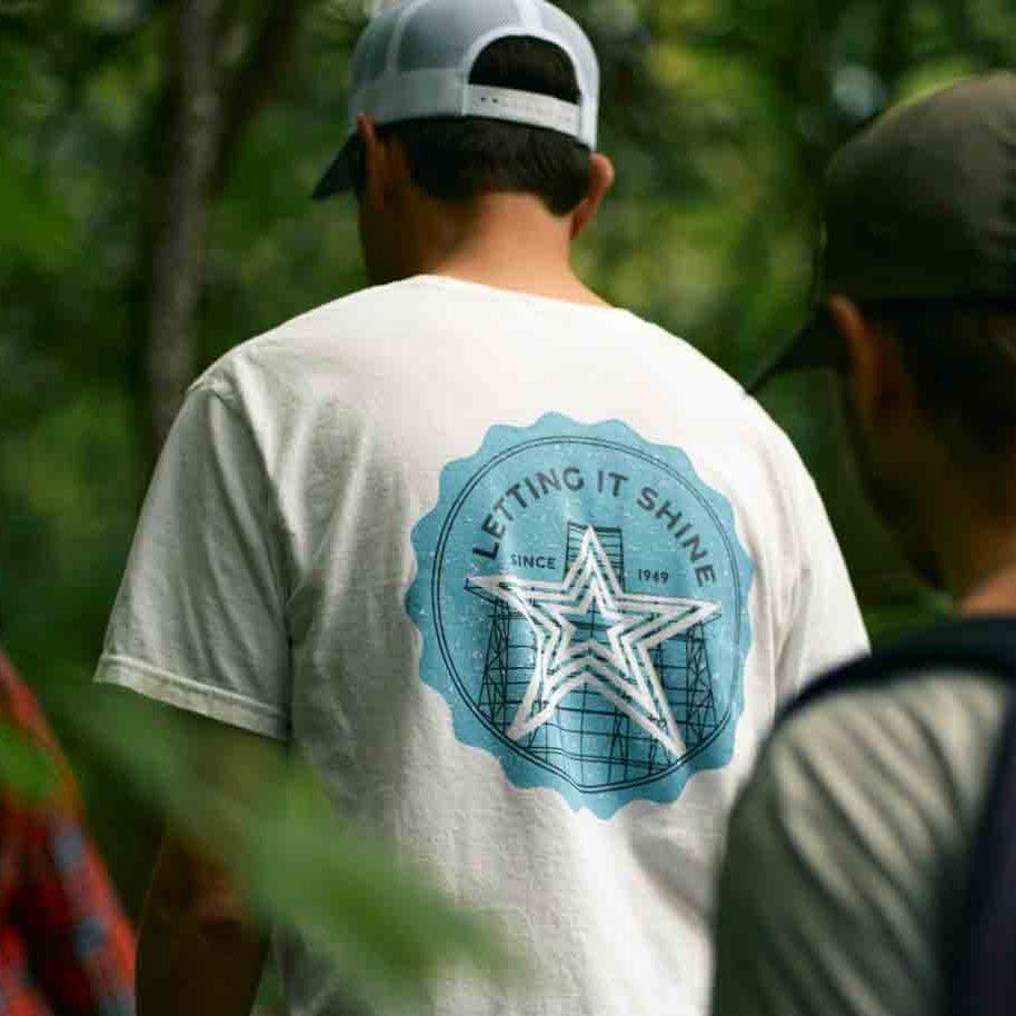 A person wearing a white "Letting It Shine" Roanoke Lifestyle t-shirt with a large blue graphic on the back featuring the Mill Mountain Star and the text "Letting It Shine Since 1949." The person is hiking outdoors, with trees and greenery in the background, highlighting the t-shirt's tribute to Roanoke's iconic landmark.
