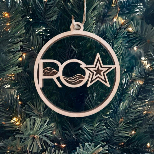 A wooden Christmas ornament by Roanoke Lifestyle, featuring a circular design with the letters "RO" and a star. The ornament is crafted with cut-out details, including a wave and star symbol on the right side. It hangs on a green Christmas tree adorned with warm white lights, adding a touch of festive charm and local pride.
